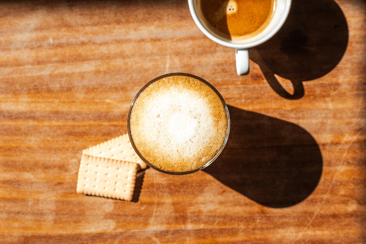 Flat lay of a coffee cup with frothy milk foam and crackers on a rustic wooden table, capturing a warm morning moment.